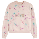 A light pink crewneck sweatshirt with a colorful paint splatter pattern in pink, purple, and blue, featuring the word 'LOVE' spelled out in rhinestones across the chest.