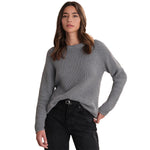 A woman wearing a gray ribbed knit crewneck sweater and black jeans, standing against a plain white background.