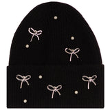 A black ribbed knit beanie hat decorated with five silver bow charms and five small white pearl-like beads, shown against a plain white background.