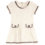 An off-white textured tweed dress with black and white fringed trim, featuring short sleeves, a round neckline, and two decorative patch pockets with button details.