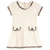 An off-white textured tweed dress with black and white fringed trim, featuring short sleeves, a round neckline, and two decorative patch pockets with button details.