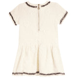 An off-white short-sleeve dress with a textured floral jacquard pattern, featuring dark brown and gold sequined trim along the neckline, sleeve cuffs, and hem, and a central gold zipper.