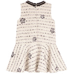 A sleeveless cream-colored dress with a black musical staff pattern and embroidered rose motifs, featuring a ruffled peplum hem and a black lace-trimmed neckline.