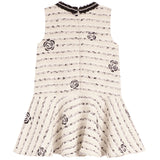 A sleeveless cream-colored dress with a black musical staff pattern and embroidered rose motifs, featuring a ruffled peplum hem and a black lace-trimmed neckline.