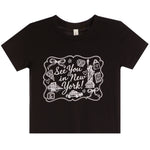 A black short-sleeve crewneck t-shirt with a white graphic print on the front that reads 'See You in New York!' surrounded by New York-themed illustrations like the Statue of Liberty, a cab, and a pizza slice.