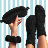 A cozy black fleece sleep set featuring a plush eye mask, matching scrunchie wristband, and fuzzy socks, shown on a person's hand and feet against a light blue and white striped background.