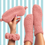 A cozy pink fuzzy sleepwear set shown on a model, including a plush eye mask held in a hand, matching fuzzy socks on the feet, and a scrunchie wristband, against a light blue and white striped background.