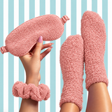 A cozy pink fuzzy sleepwear set shown on a model, including a plush eye mask held in a hand, matching fuzzy socks on the feet, and a scrunchie wristband, against a light blue and white striped background.