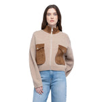 A woman modeling a cropped knit zip-up jacket in beige with brown suede-like collar, pocket flaps, and zipper trim, paired with light blue jeans against a plain white background.