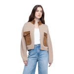 A woman modeling a cropped beige knit cardigan with brown suede-like flap pockets and a brown collar, worn over a white top and blue jeans against a plain white background.