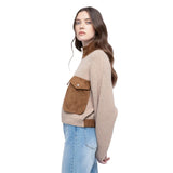 A woman wearing a beige knit sweater with brown suede-like contrast panels and a large front flap pocket, paired with light blue jeans, photographed against a plain white background.