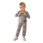 A young girl with blonde hair stands against a white background wearing a gray two-piece set with pink bow and heart appliques. She is posing with her hands forming a heart shape.