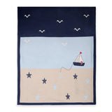 A square navy blue blanket or throw featuring a nautical design with a light blue sky, beige sand, a small sailboat, seagulls, anchors, and starfish.