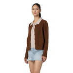 A woman modeling a brown knit cardigan with a beige collar over a light beige top and a light blue denim mini skirt, set against a plain white background.