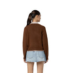 A woman shown from the back wearing a brown knit sweater over a light beige collared shirt and a light blue denim mini skirt, set against a plain white background.