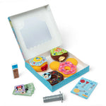 A children's toy play set featuring a light blue box with a clear window, filled with colorful donut-shaped toys, plus small accessory cards, a small container of sprinkles, and a gray spool-like piece on a white background.