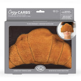 Cozy CARBS hot and cold eye pillow by Fred in lavender scent, shown inside its gray retail packaging box with a photo of a person wearing the plush croissant-shaped pillow.