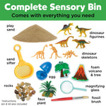 Dinosaur dig sensory bin toy set with a white plastic bin filled with sand, including dinosaur figures, a small volcano, rocks, a magnifying glass, a small brush, and a yellow scoop. The set is shown with its green storage container 