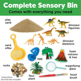 Dinosaur dig sensory bin toy set with a white plastic bin filled with sand, including dinosaur figures, a small volcano, rocks, a magnifying glass, a small brush, and a yellow scoop. The set is shown with its green storage container 