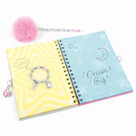 A spiral-bound notebook shown open on a white background, with a pink pom-pom pen and a beaded charm bracelet placed around it. The notebook cover is split into two halves: one side is yellow with wavy lines and the word 'YAY!', and the other is light blue with stars, a crescent moon, and the phrase 'Dream Big'.