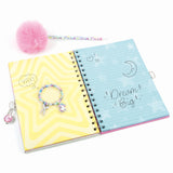 A spiral-bound notebook shown open on a white background, with a pink pom-pom pen and a beaded charm bracelet placed around it. The notebook cover is split into two halves: one side is yellow with wavy lines and the word 'YAY!', and the other is light blue with stars, a crescent moon, and the phrase 'Dream Big'.
