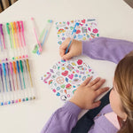A child's hands and arms are shown writing on a sheet of colorful sticker-like graphics while sitting at a white table. Several colored gel pens are arranged in clear plastic cases on both sides of the table.