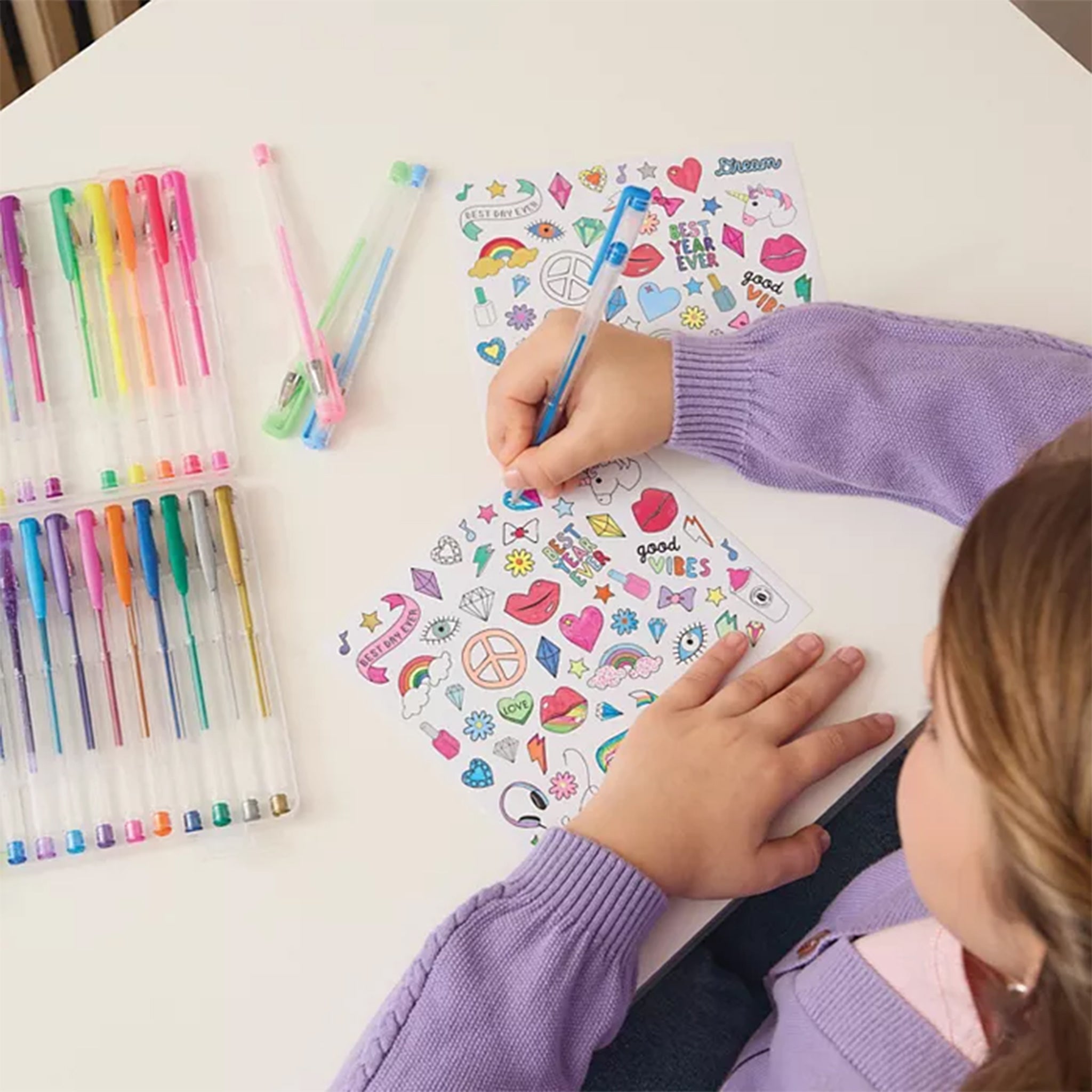 A child's hands and arms are shown writing on a sheet of colorful sticker-like graphics while sitting at a white table. Several colored gel pens are arranged in clear plastic cases on both sides of the table.