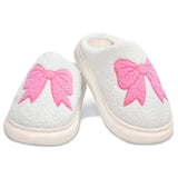 A pair of white fuzzy slip-on slippers with large pink bow designs on the top, shown on a clean white background.