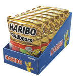 A display box filled with multiple foil packets of Haribo Goldbears candy on a plain white background. The packets are metallic gold with the Haribo brand and product name clearly visible, and the display box is blue with Haribo branding and cartoon bear illustrations.