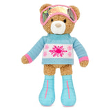 A plush teddy bear dressed in ski attire, featuring a light blue knit sweater with a pink snowflake pattern, matching blue ski boots, pink-trimmed gloves, and reflective pink goggles, set against a plain white background.