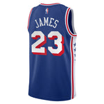 A blue basketball jersey shown from the back, featuring 'JAMES' and the number '23' in white with red outlines. The jersey has red and white trim on the shoulders and sides, and an NBA logo is visible at the top collar.