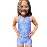 A young girl with long brown hair and clear swim goggles is wearing a matching two-piece swimsuit with a blue galaxy print and pink peace signs, flowers, rainbows, etc., set against a plain white background.