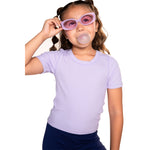 A young girl with braided hair wearing a light lavender short-sleeve crewneck t-shirt and dark navy pants, holding a pair of pinkish-purple sunglasses to her face while blowing a bubble gum bubble, set against a plain white background.