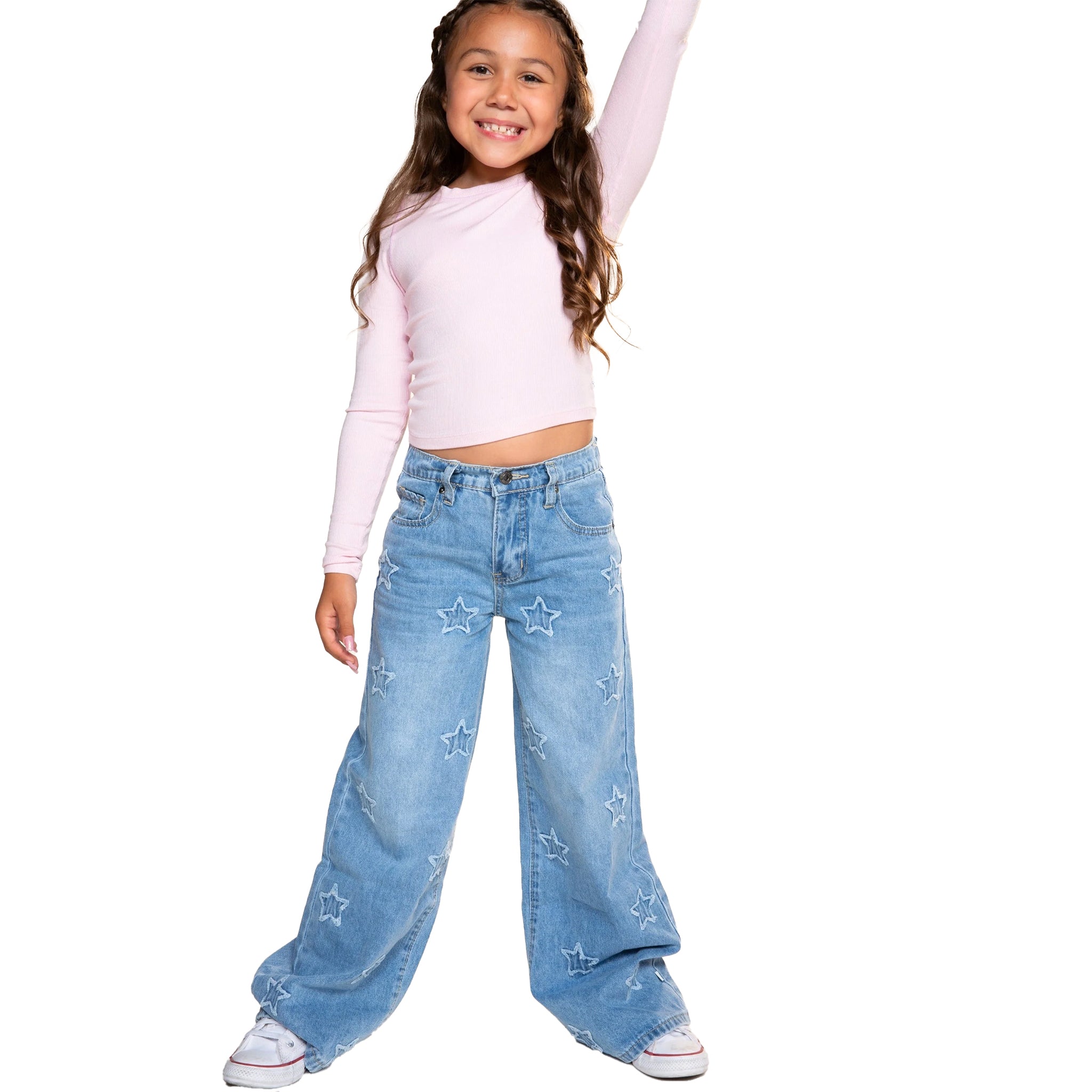 A young girl with long brown hair stands against a white background wearing a light pink long-sleeve crop top and light blue wide-leg denim jeans decorated with white star prints.