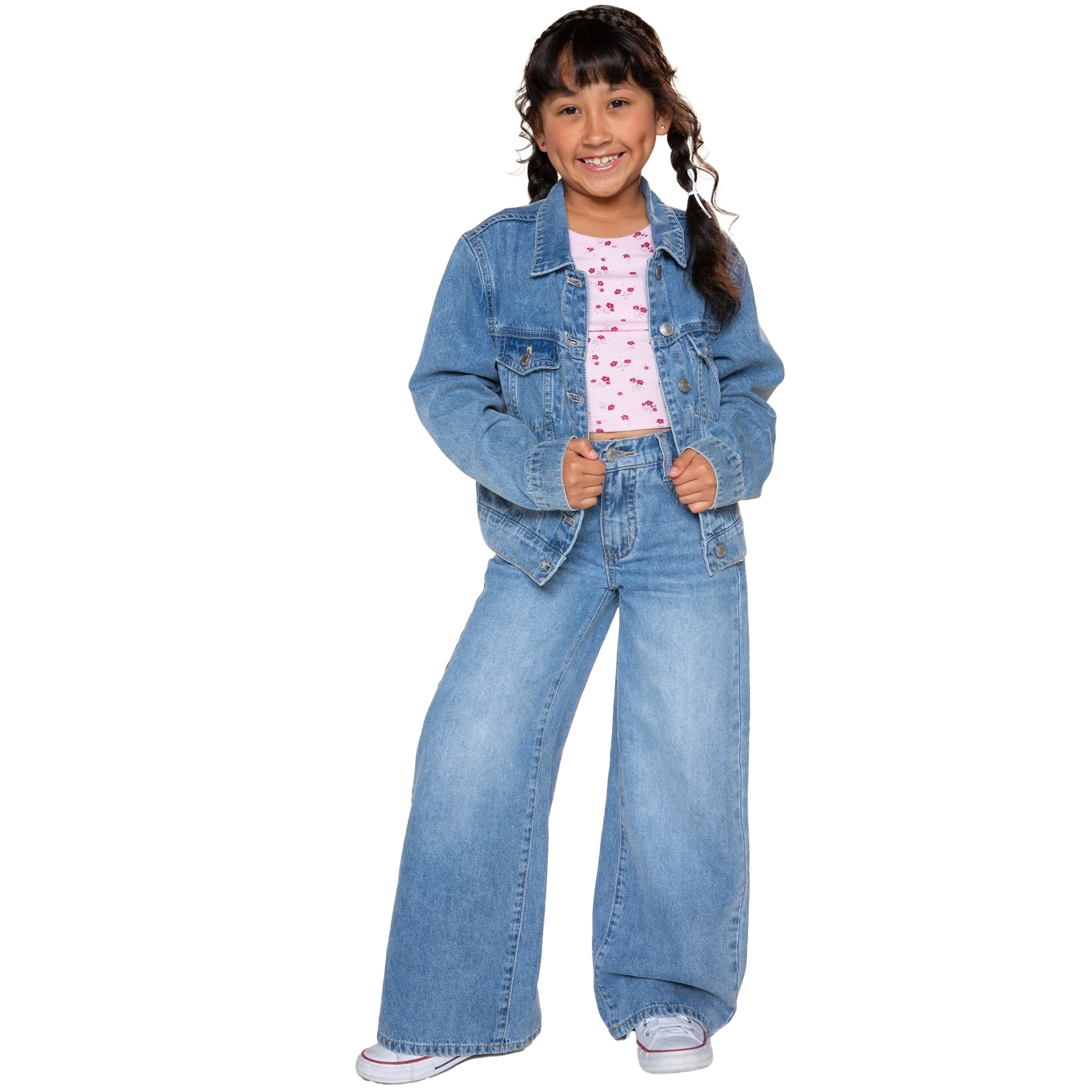 A young girl with dark hair in pigtails stands against a white background wearing a medium blue denim jacket and matching wide-leg jeans over a pink patterned t-shirt.