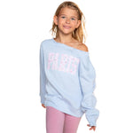 A young girl with blonde hair wearing a light blue off-the-shoulder sweatshirt with 'PARIS' printed in pink and white on the front, paired with pink leggings, against a solid white background.