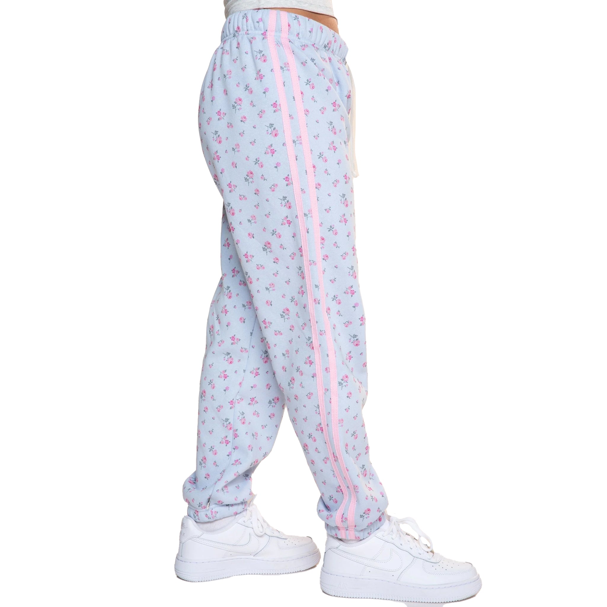 Light blue jogger sweatpants with a pink floral print, featuring a pink stripe down the side and white sneakers shown on a model from the waist down against a white background.