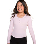 A young girl with long dark hair smiling, wearing a light pink long-sleeve henley shirt with three buttons and black pants, set against a plain white background.
