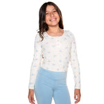 A young girl smiling, wearing a long-sleeve white henley-style top with a light blue floral pattern and light blue leggings, set against a plain white background.