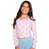A young girl with long brown hair smiling, wearing a light pink long-sleeve crop top with a small pink and purple floral pattern, paired with light blue leggings, against a solid white background.