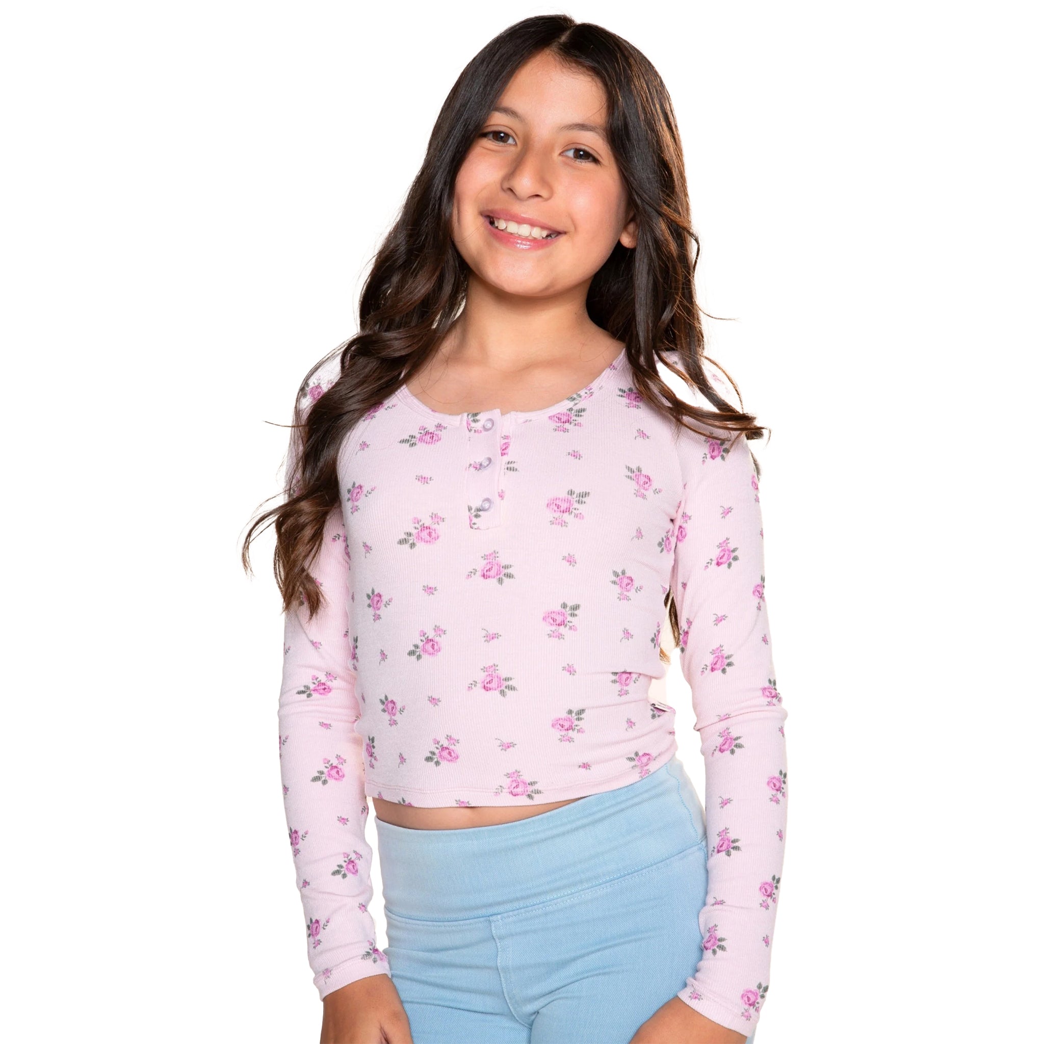 A young girl with long brown hair smiling, wearing a light pink long-sleeve crop top with a small pink and purple floral pattern, paired with light blue leggings, against a solid white background.