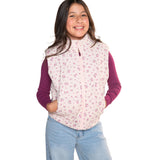 A young girl smiling and wearing a light pink zippered fleece vest with a small purple floral pattern, layered over a dark purple long-sleeve shirt and paired with light blue jeans, set against a plain white background.