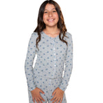 A young girl smiling and wearing a light gray long-sleeve pajama top with a blue floral pattern and matching pants, photographed against a solid white background.