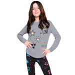 A young girl with long brown hair stands against a white background wearing a heather gray long-sleeve shirt with colorful graphic prints (star, heart, rainbow, 'LOVE' text) and black leggings with a colorful space pattern.