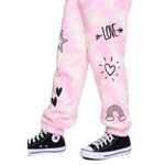 Pink and white tie-dye jogger pants with black graphic prints including hearts, a rainbow, a star, and the word 'LOVE' with arrows, shown on a child's legs with black Converse sneakers against a white background.