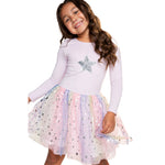 A young girl smiling and wearing a light lavender long-sleeve top with a silver sequined star on the chest, paired with a fluffy pastel tulle skirt covered in silver star sequins, set against a plain white background.