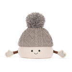 A cute, smiling marshmellow knit pom-pom hat with a gray cable-knit top section, a cream-colored bottom section, and small brown arms ending in cream mittens, shown on a solid white background.