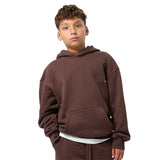A child model wearing a matching dark brown sweatshirt and jogger pants set, styled with a white undershirt and tan shoes, standing against a light gray studio background.