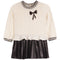 A girls' long-sleeve knit top and pleated skirt set in cream and black. The cream cable-knit top features a striped crew neck, striped cuffs, a ruffled hem, and a black sequined bow on the chest.