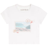 A white short-sleeve crewneck t-shirt by Katie J, featuring a vintage postage stamp graphic on the front with a coastal landscape, shells, and a stamp cancellation mark.